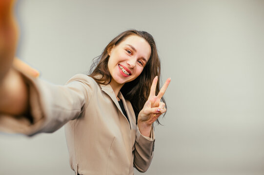 Smiling lady in beige jacket taking selfie on camera smartphone on beige background, looking at camera with happy face.