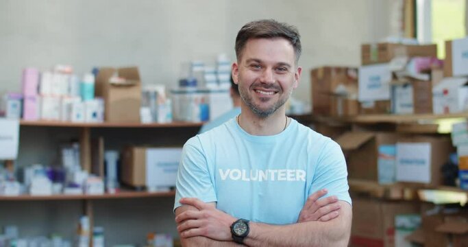 Caucasian food bank worker in blue uniform keeping arms crossed and smiling while standing among shelves with donations. Portrait of positive male volunteer.