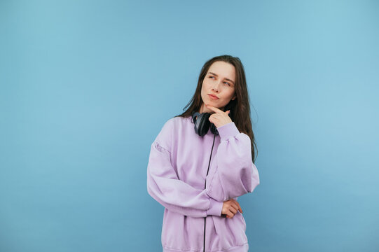 Portrait of beautiful pensive woman in purple sweatshirt on blue background, looks aside at copy space and thinks