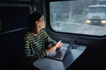 Attractive woman in wireless headphones works on a train on a laptop and uses a smartphone, looks out the window