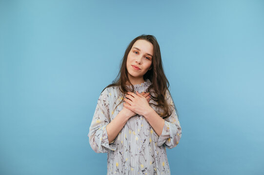 gentle lady in a dress stands on a blue background and touches her hands to her heart and looks at the camera.