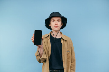 Surprised young man in panama and shirt stands with smartphone in hand on blue background and shows screen with black screen.