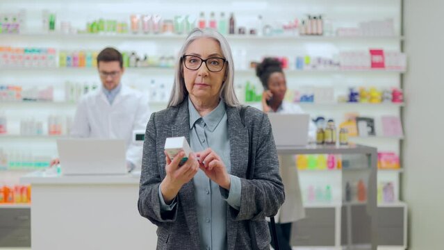 Portrait Of Beautiful Senior Woman Smiling And Looking At Camera While Standing In Drugstore With Medication In Hands. Multiracial Male And Female Pharmacists Working On Computers On Background.