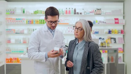 Competent male pharmacist in eyewear and lab coat standing with senior woman in drugstore and advising medication according disease symptoms. Female pensioner buying pills at pharmacy. - Powered by Adobe