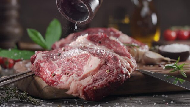 Two raw rib eye steaks with spices and herbs on a wooden kitchen table, sprinkle with sea salt on top. 
