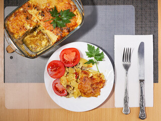 Portion of potato casserole with meat on plate with fresh tomatoes, cutlery on table 