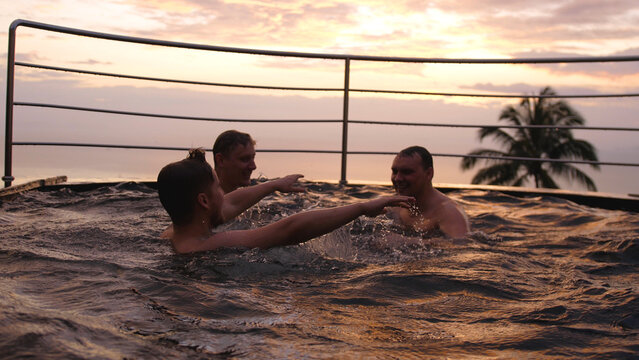 Happy friends swimming in pool splashing playfully enjoying summer vacation having fun celebrating summertime holiday at luxury hotel with ocean view sunset