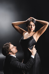 Man in suit touching waist of sexy girlfriend in silk dress on black background.