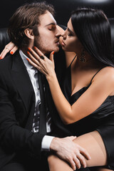 Side view of sexy woman in dress touching boyfriend in suit on couch on black background.