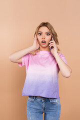 thrilled woman touching face while talking on smartphone isolated on beige.