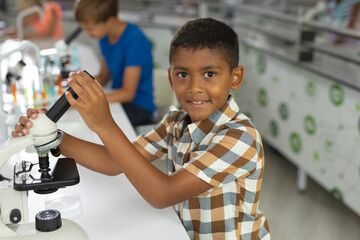Portrait of biracial elementary schoolboy holding microscope while sitting at desk in laboratory