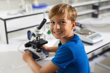 Portrait of smiling caucasian elementary schoolboy with microscope sitting at desk in laboratory - obrazy, fototapety, plakaty