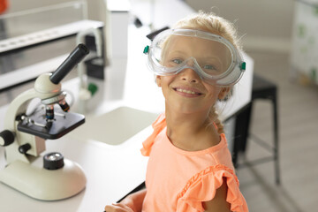 Portrait of smiling caucasian elementary schoolgirl with protective glasses sitting in laboratory