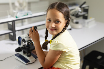 Portrait of smiling caucasian elementary schoolgirl with microscope sitting at desk in laboratory