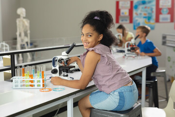 Portrait of smiling biracial elementary schoolgirl with microscope sitting at desk in laboratory