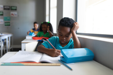 African american elementary schoolboy with head in hand writing on book at desk in classroom