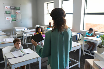 Caucasian young female teacher with digital tablet talking to multiracial elementary students