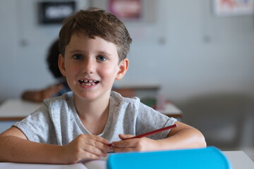 Portrait of cute smiling caucasian elementary schoolboy with pencil sitting at desk in classroom