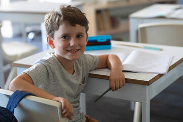 Portrait of cute smiling caucasian elementary schoolboy sitting at desk in classroom