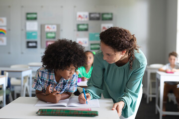Caucasian young female teacher assisting african american elementary schoolboy studying at desk