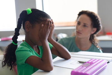 Caucasian young female teacher consoling sad african american elementary girl with covered face