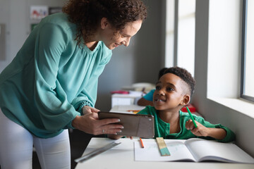 African american elementary boy looking at caucasian young female teacher with digital tablet