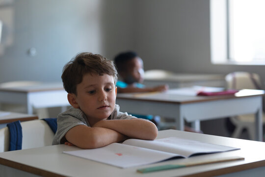 Caucasian elementary schoolboy reading book while sitting at desk in classroom