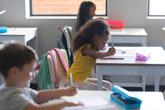 Multiracial elementary school students writing on book while studying at desk in classroom
