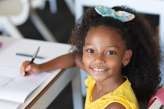 Portrait of smiling african american elementary schoolgirl writing on book at desk in classroom