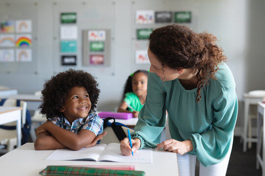 Caucasian young female teacher teaching african american elementary schoolboy studying at desk - Powered by Adobe
