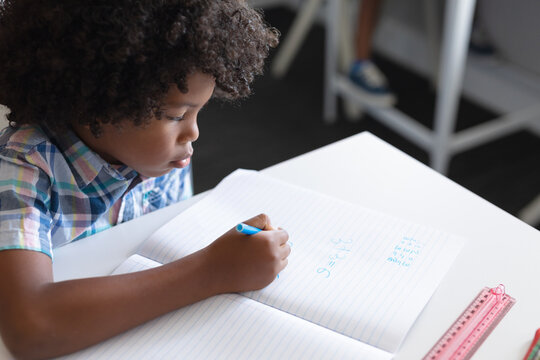 High angle view of african american elementary schoolboy writing on book at desk in classroom