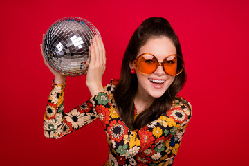 Photo of excited pretty woman wear flower print dress eyewear holding disco ball isolated red color...