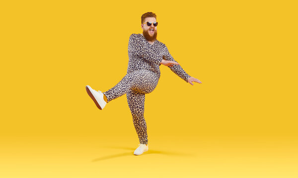 Plus size guy in funny pyjamas having fun in modern studio. Happy carefree fat man wearing comfortable leopard PJs and sunglasses dancing isolated on yellow background. Crazy party and fashion concept
