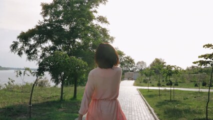 Woman in pink dress smiling walking on river bank with hair flying on wind, blue sky background with sunlight flare.