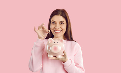 Smiling frugal woman throws coin into piggy bank which she holds in her hand on pastel pink studio...