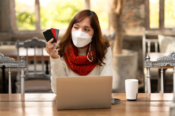 Woman sitting in cafe with white mask, shopping online by using laptop and pay by credit card, seller promotion in Winter for buyer to get cashback after payment, internet banking, eCommerce business