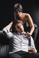 Obraz premium Sensual woman in dress taking off tie from boyfriend holding whiskey on couch isolated on black.