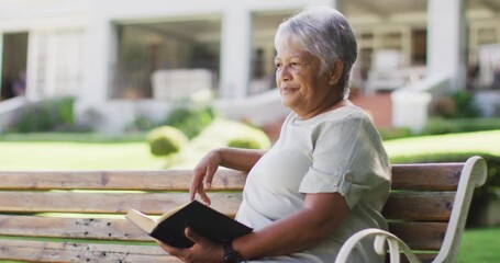 Video of relaxed biracial senior woman reading, sitting on bench in garden - Powered by Adobe