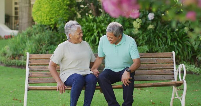 Video of happy biracial senior couple embracing and sitting on bench in garden