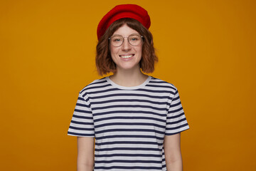 indoor portrait of young ginger female, wears stripped t shirt and glasses posing over orange background looking into camera with broad smile on her face