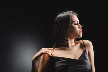 Fototapeta premium Side view of sensual brunette woman touching silk dress on black background.