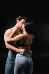 Woman in jeans and bra kissing sexy boyfriend isolated on black.