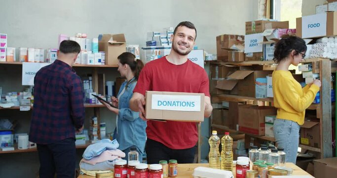 Portrait of male food bank worker in casual attire showing donation box while standing at warehouse with group of volunteers behind. Concept of people, social service and humanitarian aid.