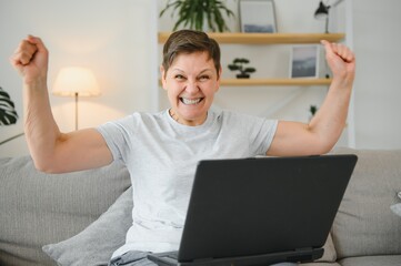 Excited mature woman looking at laptop screen, reading good news in message, celebrating online lottery win, rejoicing success, overjoyed older female sitting on couch at home, using computer.