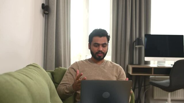 Video call concept. Indian freelancer man waving hello into laptop webcam, guy connected via video with family, friends or colleagues working remotely from home