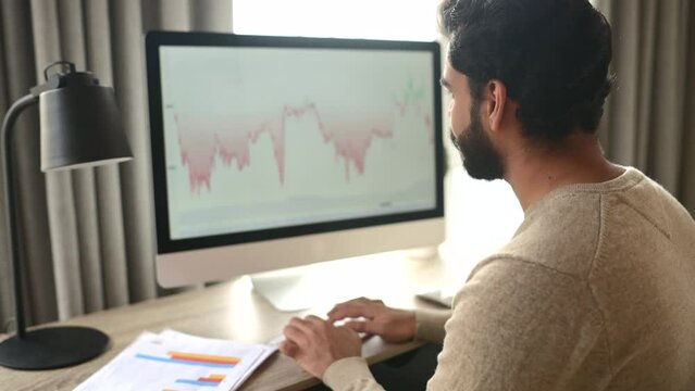 Indian male trader investor using computer for remote work, making cryptocurrency financial market analysis, trading data index chart graph on the screen, researching stock market