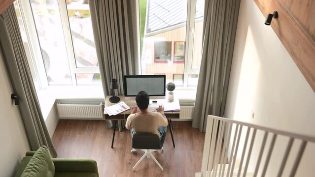 Back view of the clever and concentrated caucasian guy is doing paperwork. Man looking through paper sitting at the desk with a computer in office. Man checking report concept
