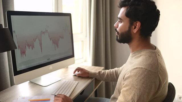 Concentrated high-skilled bearded Indian broker sitting at table and using computer to trade on stock market, sitting in home office, making stock market research