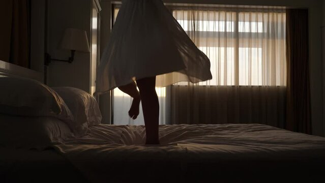 Happy woman is having fun - jumping and dancing on the bed, her skirt flying around. Low half shot, camera shows legs and skirt. Dark room, sunset sun gleam through window on background