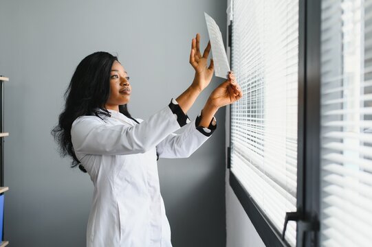 Young attractive african female doctor radiologist looking at x-ray image, while standing in light modern office in clinic